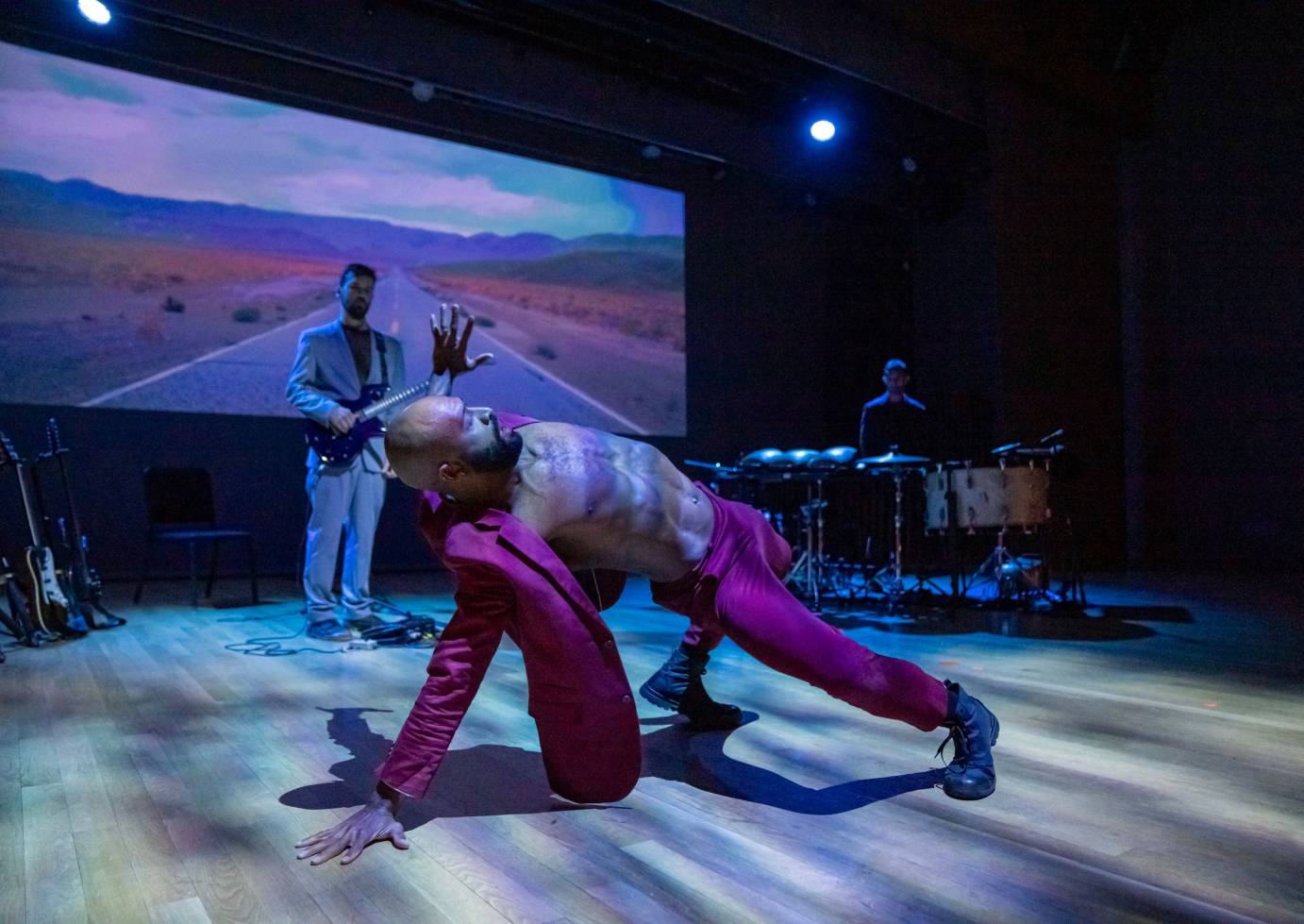 guitarist and drummer onstage in front of projection of a long winding highway road and mountains in the distance. A dancer is in the foreground in a red suit and no shirt, in a hinge with one hand on the floor