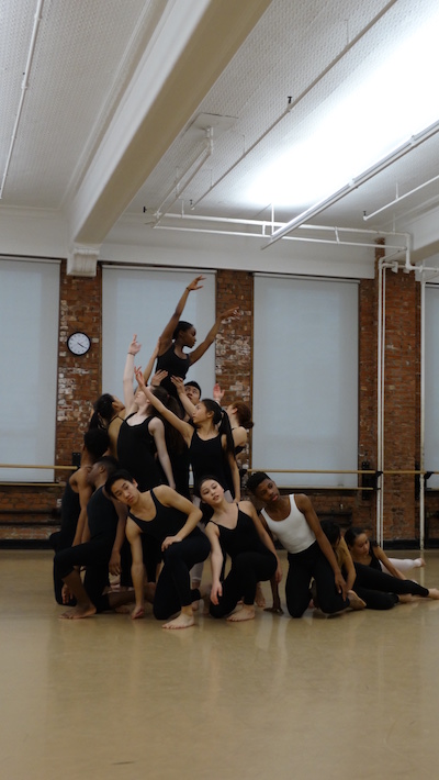 A group of students are arranged in a cascading clump with some dancers kneeling on the floor, others standing and one is lifted in the air.