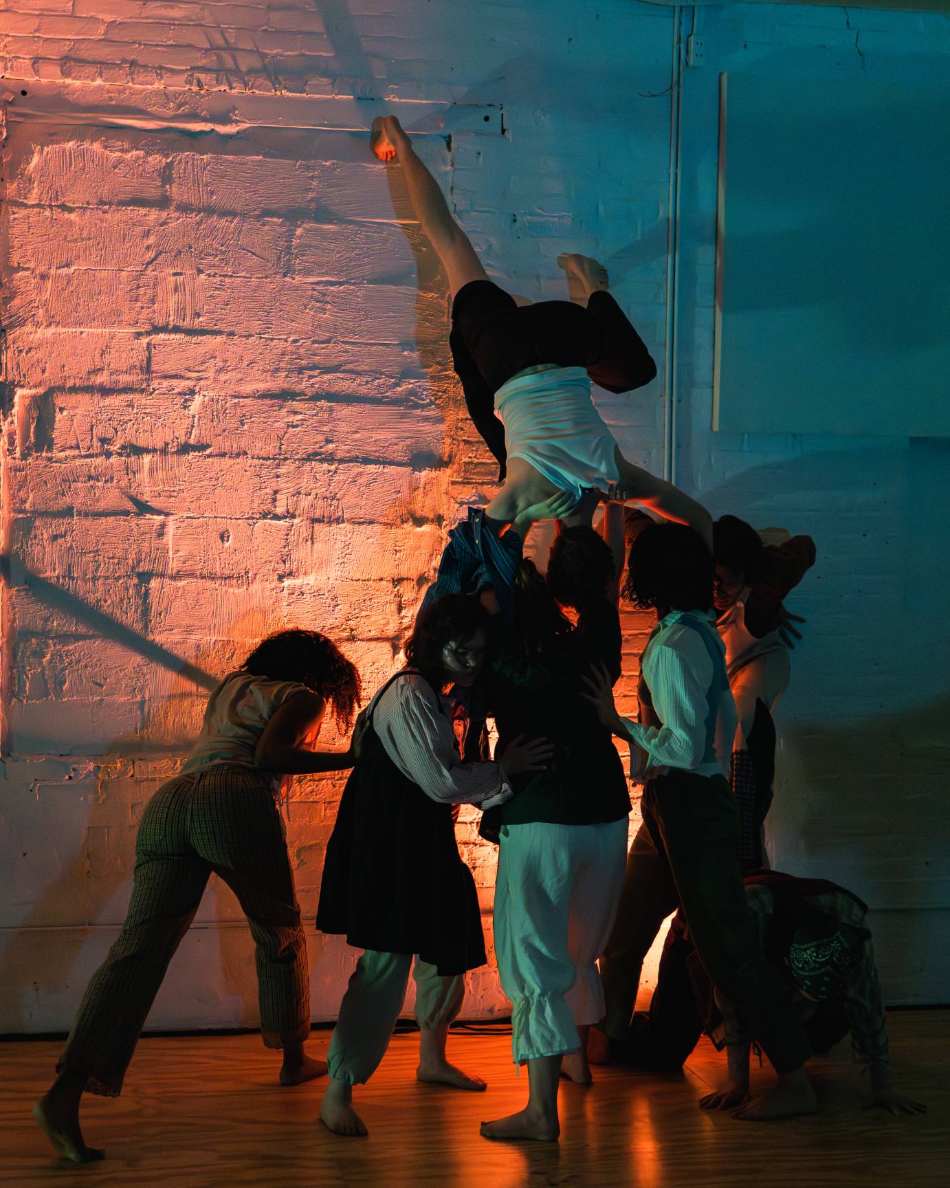 The dancers hold a dancer, wearing a sleeveless white shirt and black pants, high above them upside down.