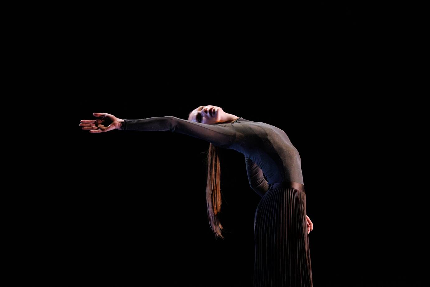 light-haired woman in grey long sleeve leotard with a black pleated skirt bends backward from her waist extending her right arm to follow her body's arch 