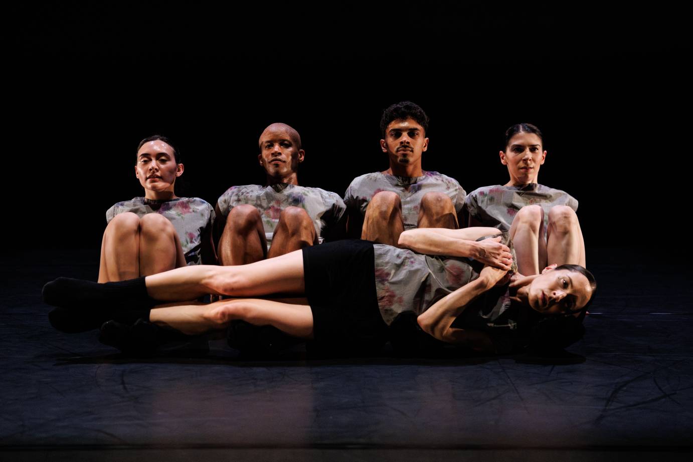 against a black background 4 dancers sit upright facking us with their knees drawn towards their chests, another dancer, lying on her side rests her entire body sideways on top of the quartets feet as she clasps her to her left shoulder