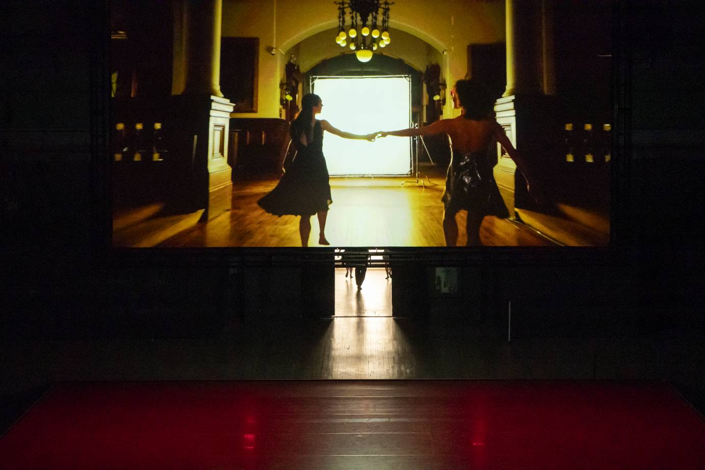 Romeo and Juliet played by two women, hold hands and run down an Armory corridor, the light from a door bathing them.