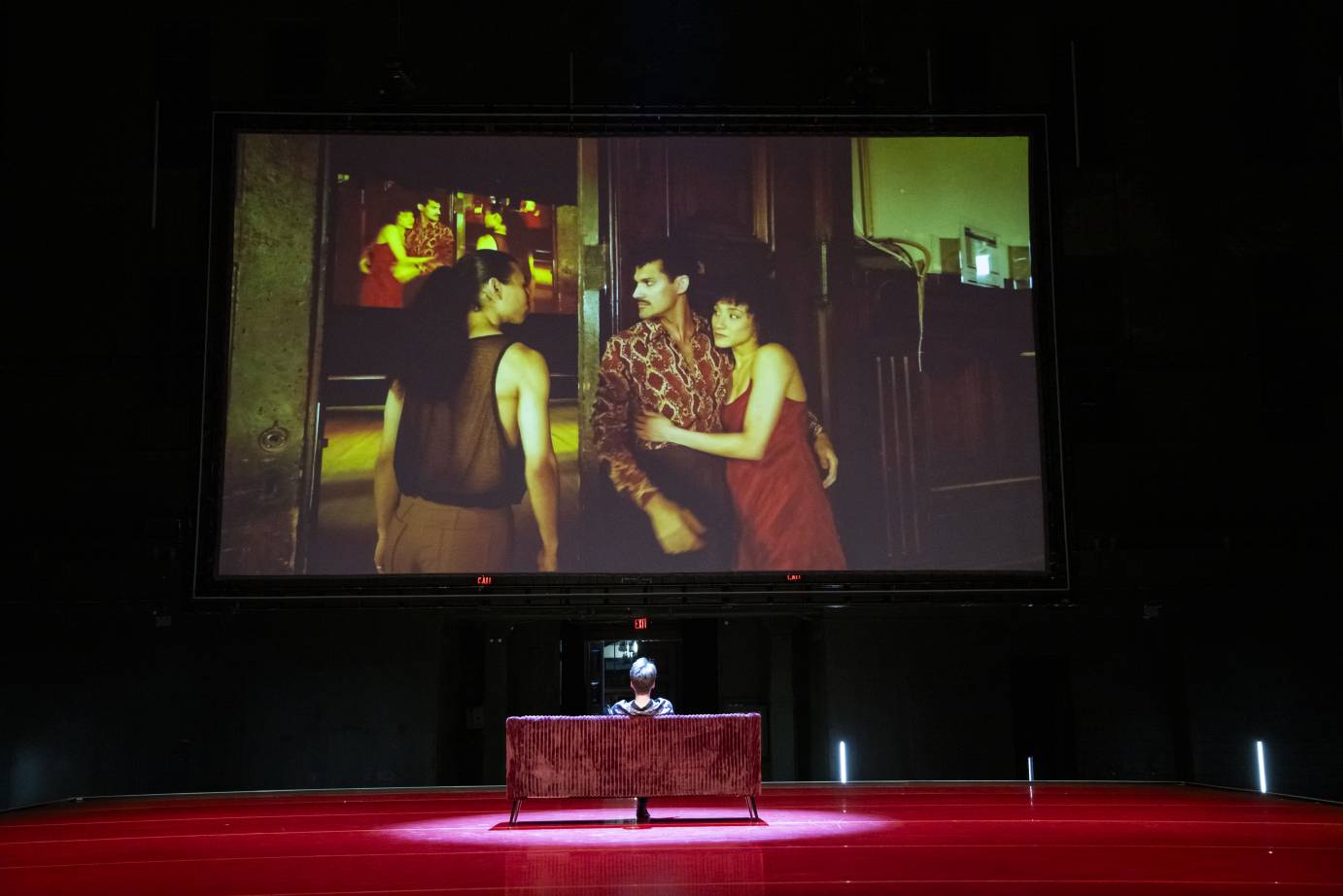 A dancer sits on a red sofa facing action on the big screen: a couple is confronted by a third performer.