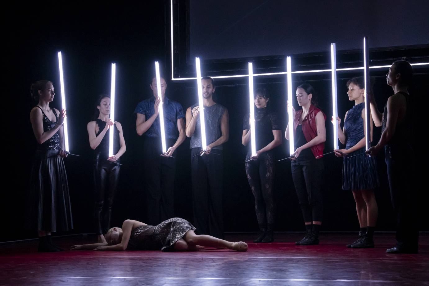Seven dancers, standing side by side, hold long neon 'sticks' in front of their bodies. Juliet lies on her side on the floor in front of them.