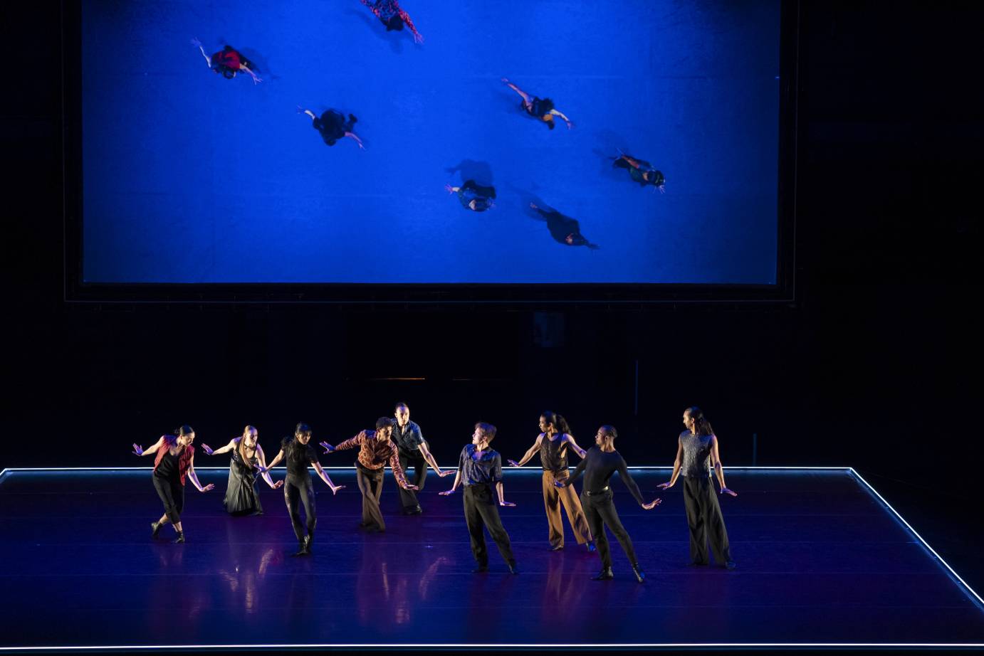 The ensemble dancers on stage are seen from above on screen bathed in blue.