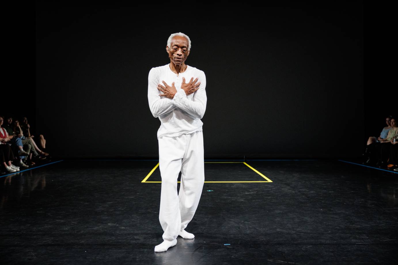A white haired dancer arms crossed over the chest dressed in a long-sleeved top, white pants and white socks.