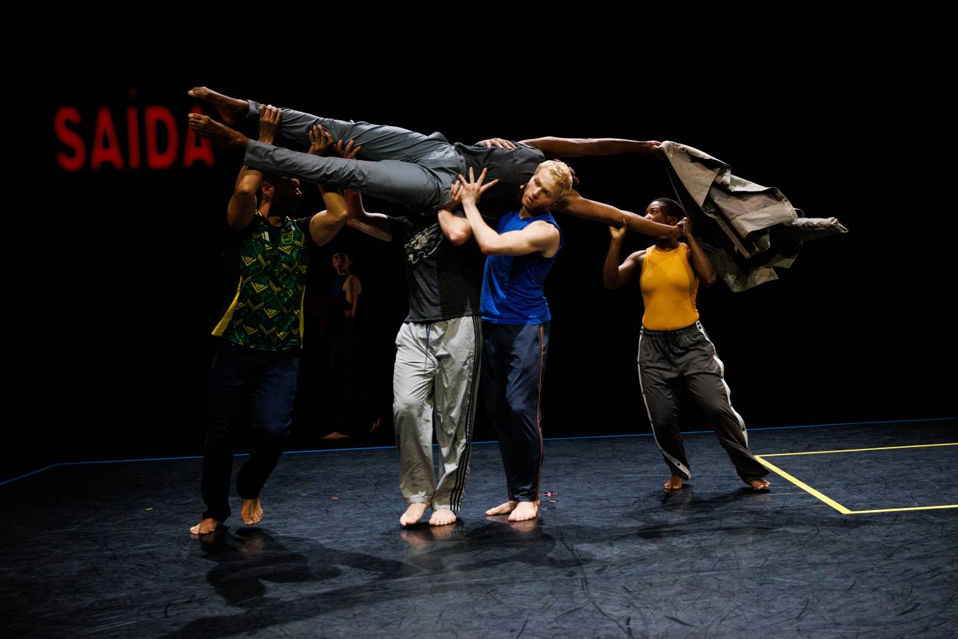 A group of dancers in pedestrian clothing lifting a man on their shoulders. His arms are flung overhead.