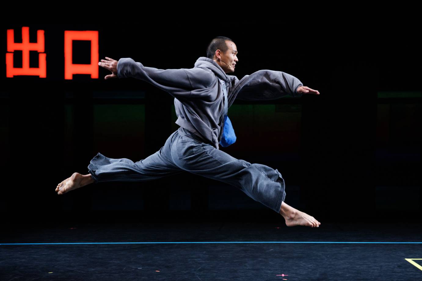 A smiling male dancer in profile in a bent leg leap passing red Chinese-like writing.
