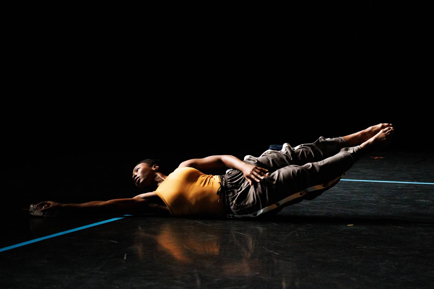 A dancer costumed in an orange sleeveless top and loose gray bottoms dives to the edge of the space that is ringed in blue tape.