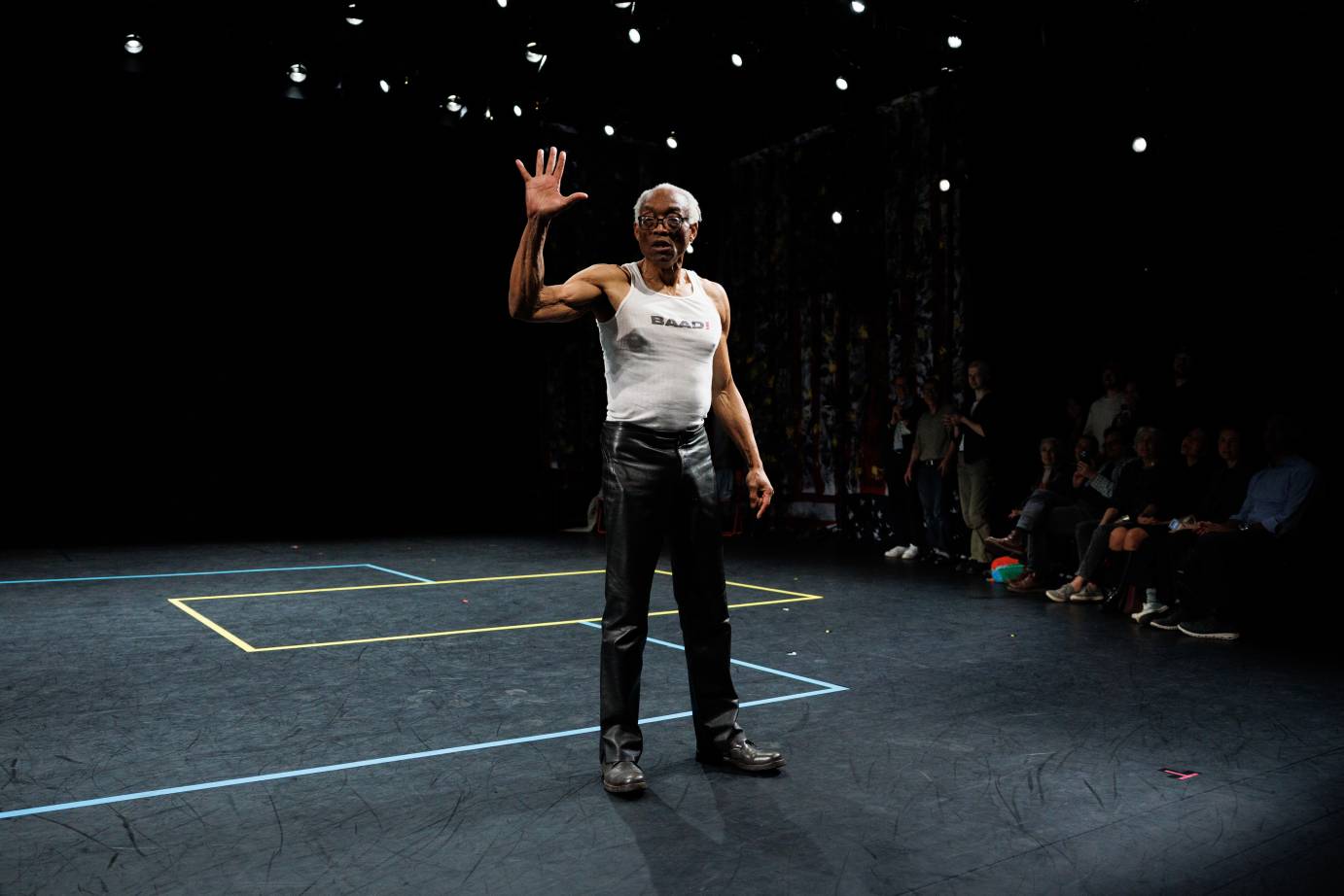 A white-haired man in a white sport tank top, black pants and boots, lifts his arm with palm to audience fingers splayed.