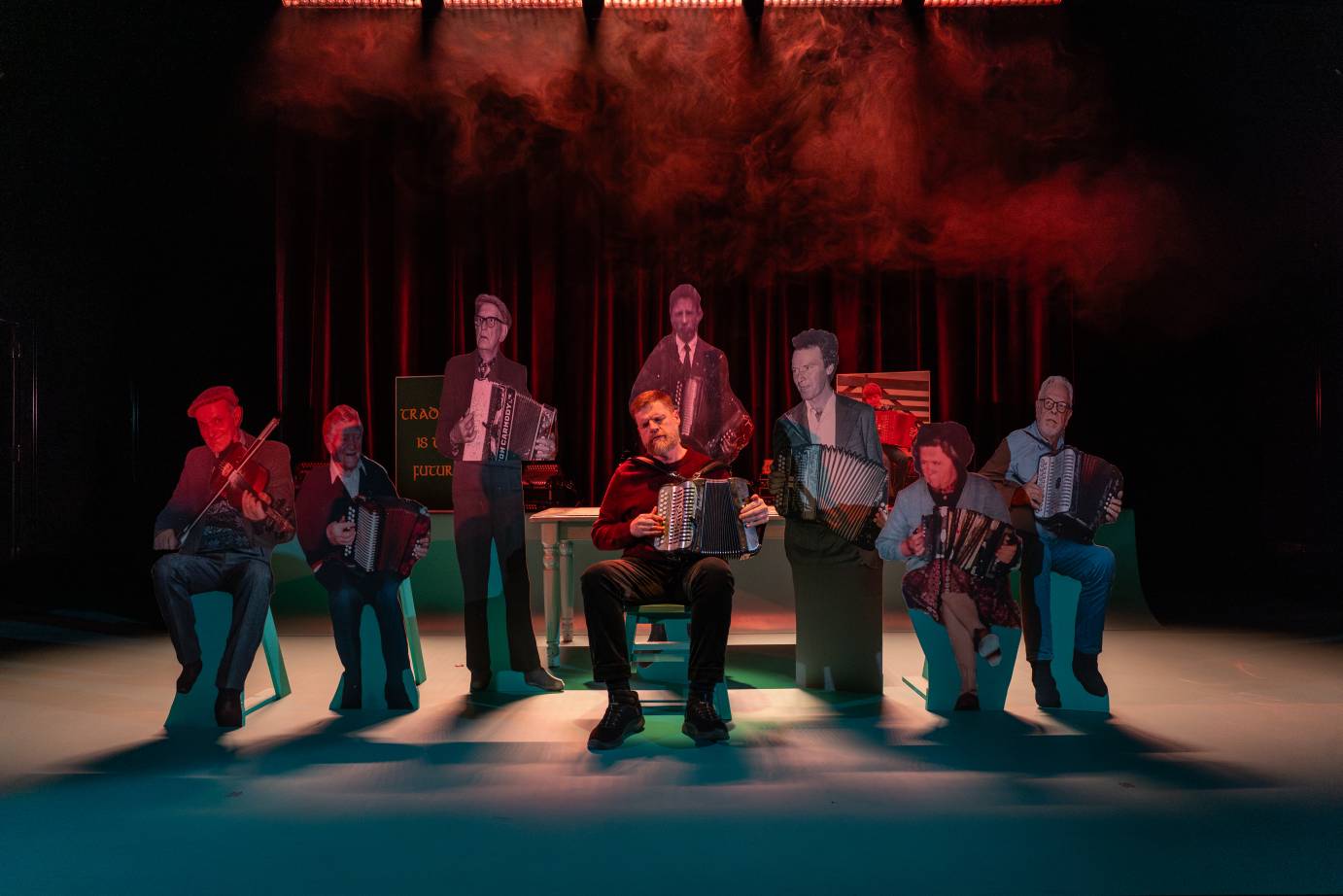 The seated accordianist is surrounded with life like cut out standing and sitting photos of the accordianist mentors.