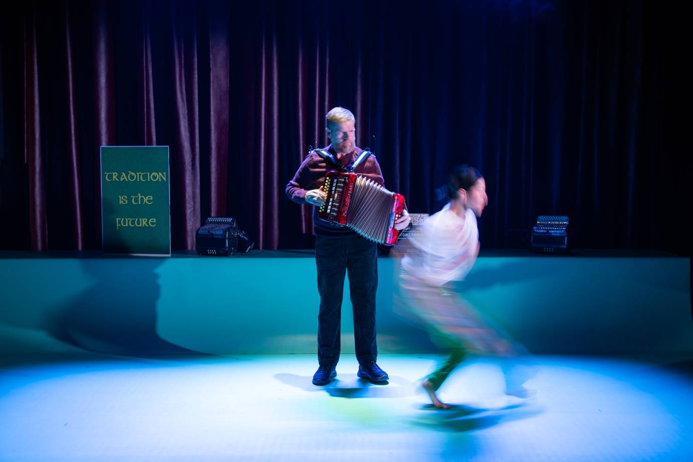 An accordianist watches a woman dancer in white. Her image is blurred.