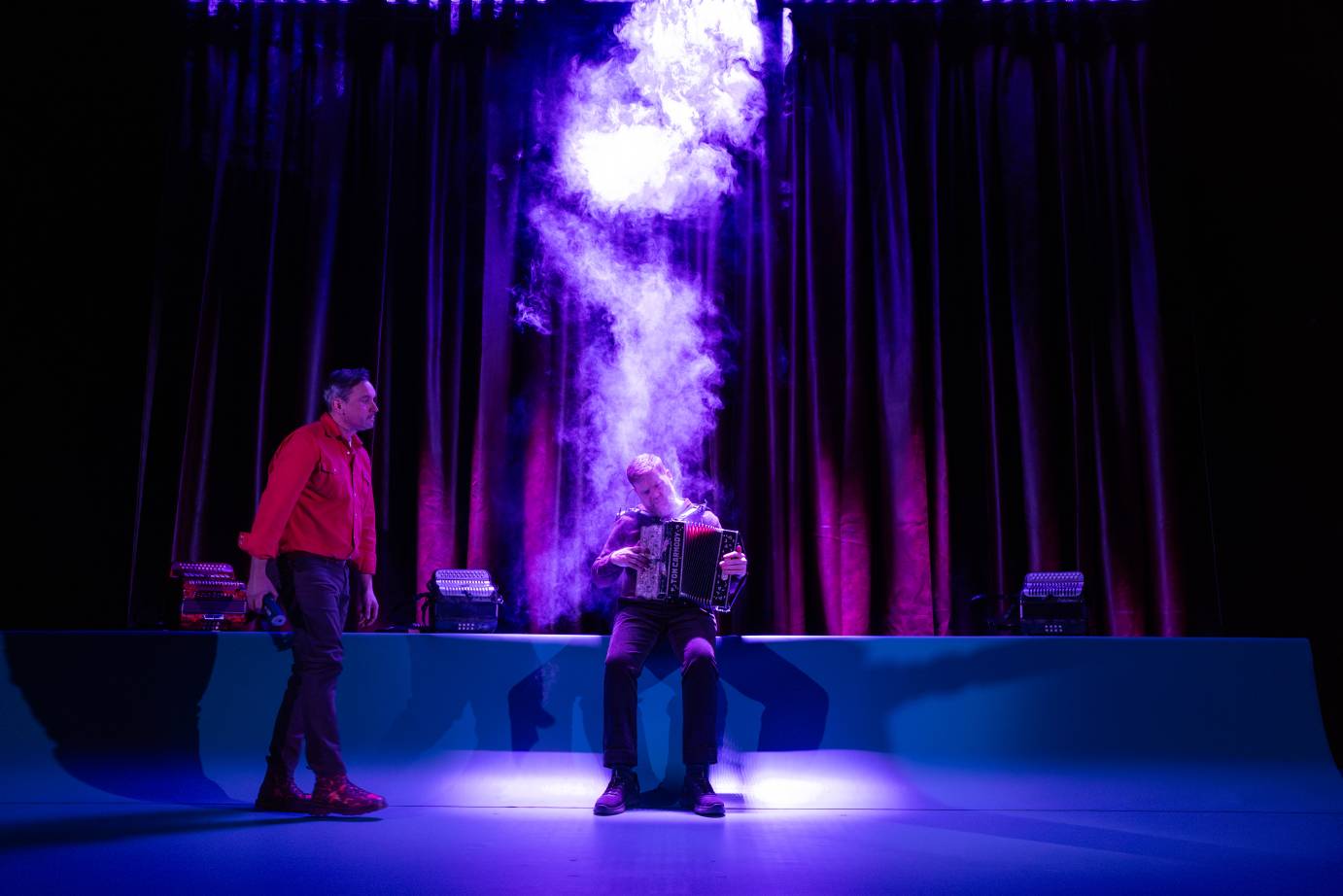 A man wearing a red shirt stands to the side of the seated accordianist. A flame of purple smoke shoots up behind the accordianist.