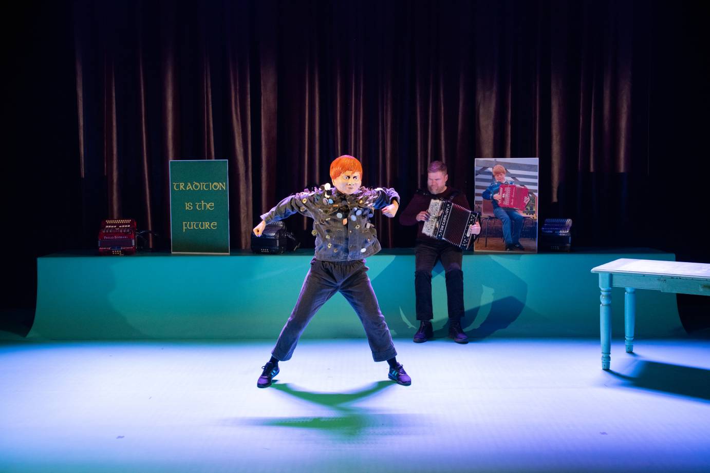The dancer, wearing a red-headed mask, and a jacket with dangling pins, stands with her legs apart. The accordianist accompanies behind her.
