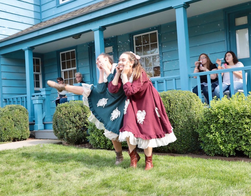 Two women smile with their hands framing their faces. People watch behind them on blue wrap-around porch.