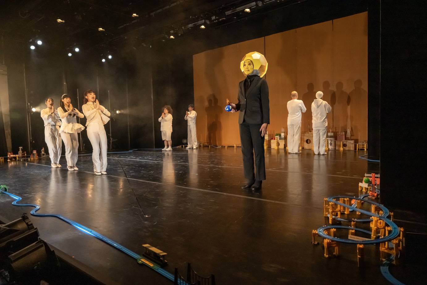 Seven performers in white are arranged on stage according to a performer dressed in a black suit wearing a moon mask. Three women are in a line to the right of the moon figure while two duets are upstage. All their hands clasp in a prayer position.
