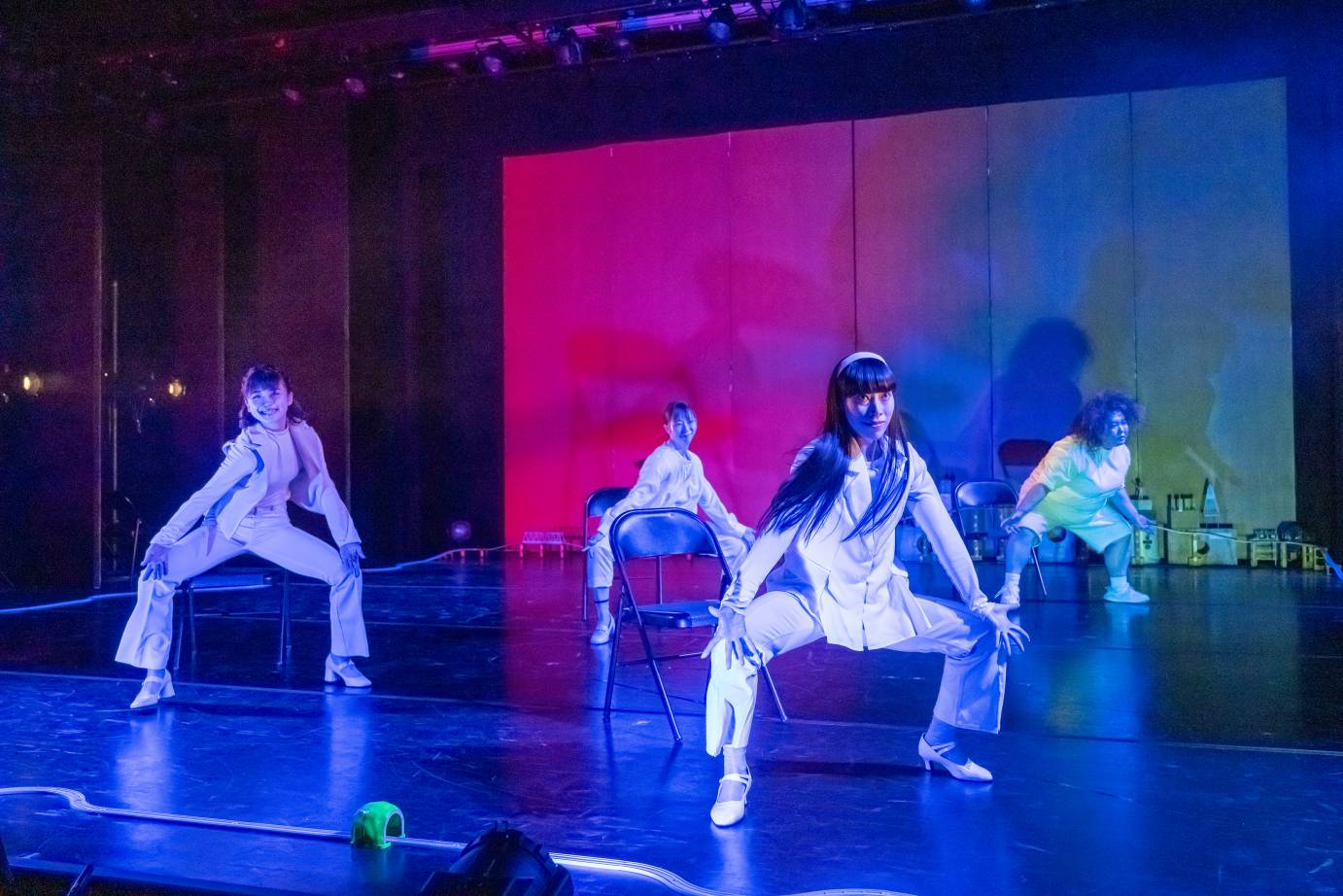 The four women in white rise out of their chairs with legs spread in 2nd position and smiles on their faces. The backdrop moves from red to blue to green.