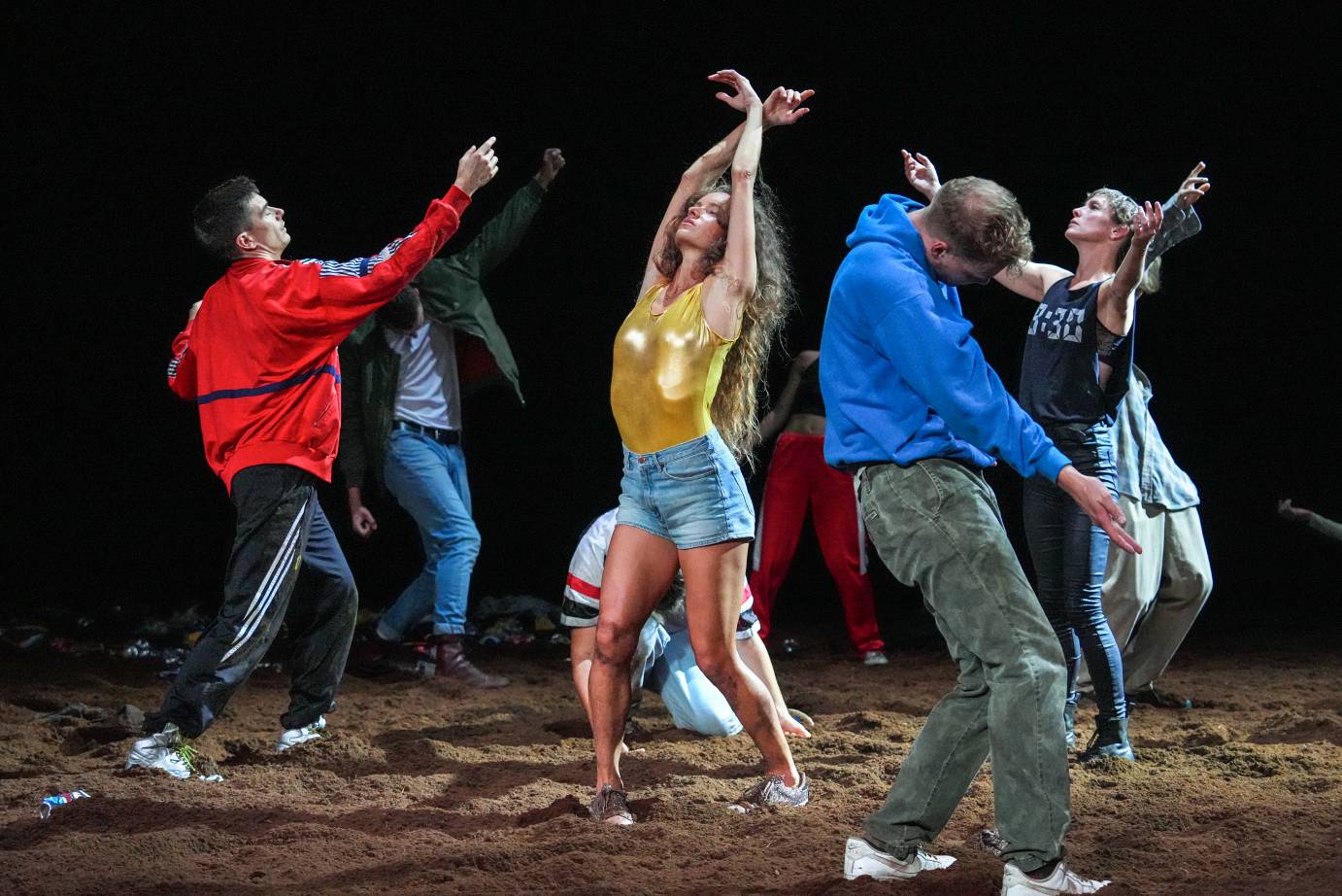 on a floor of dirt a group of revelers in casual clothes dance as if lost in an ecstatic stupor. the central revelr has long wavey brown hair, wears a golden leotard, hot pants and silver sneakers