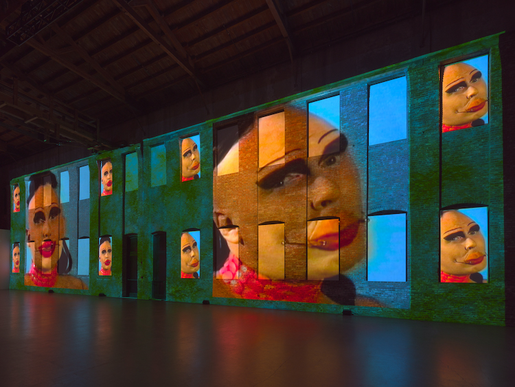 projected face Large with several smaller versions of performance artist with dark made up eyes and full red lips with lower lips pierced by large safetly pins running from each cheek to lower lip