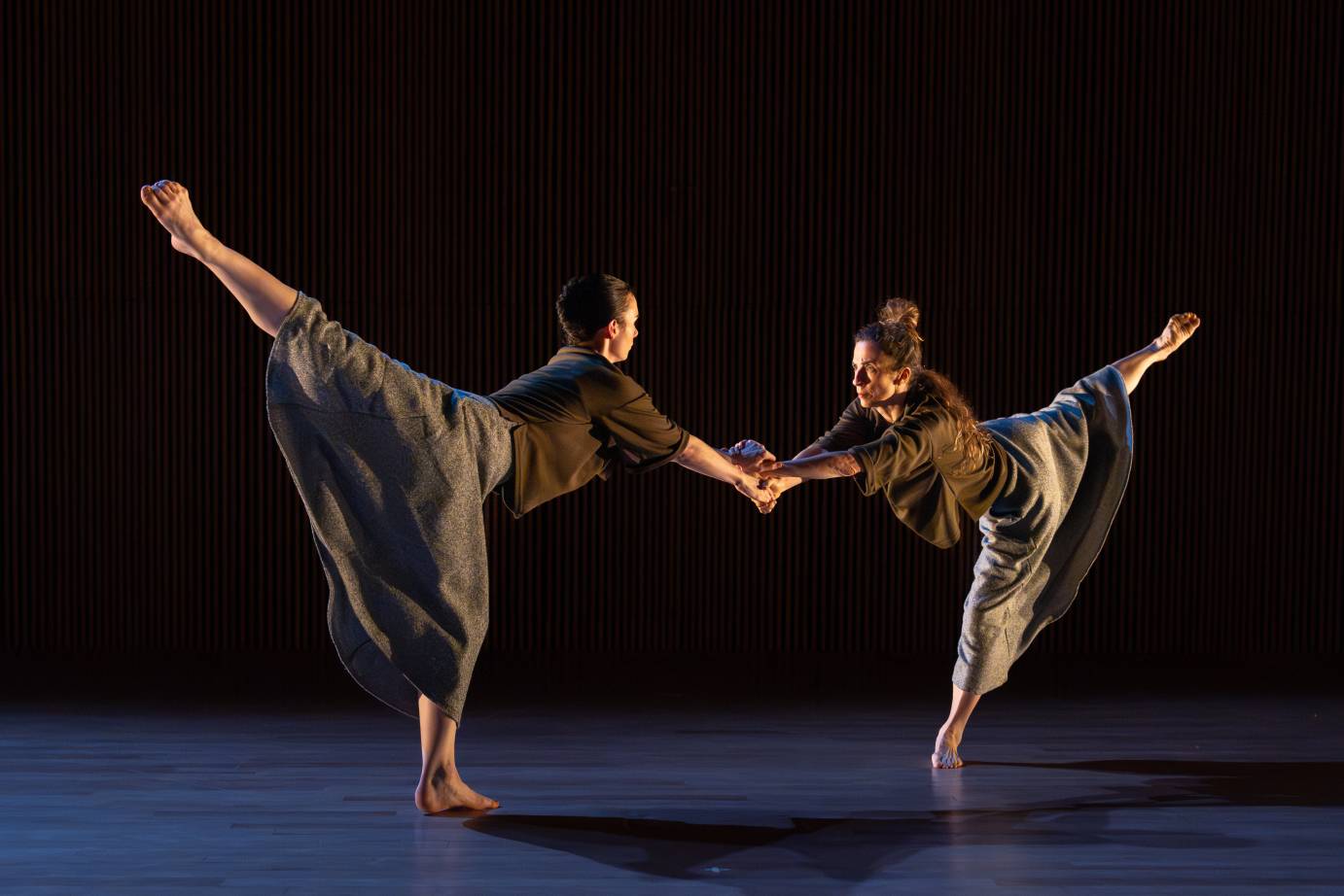 Two women in high arabesques face one another clasping and crossing wrists. 