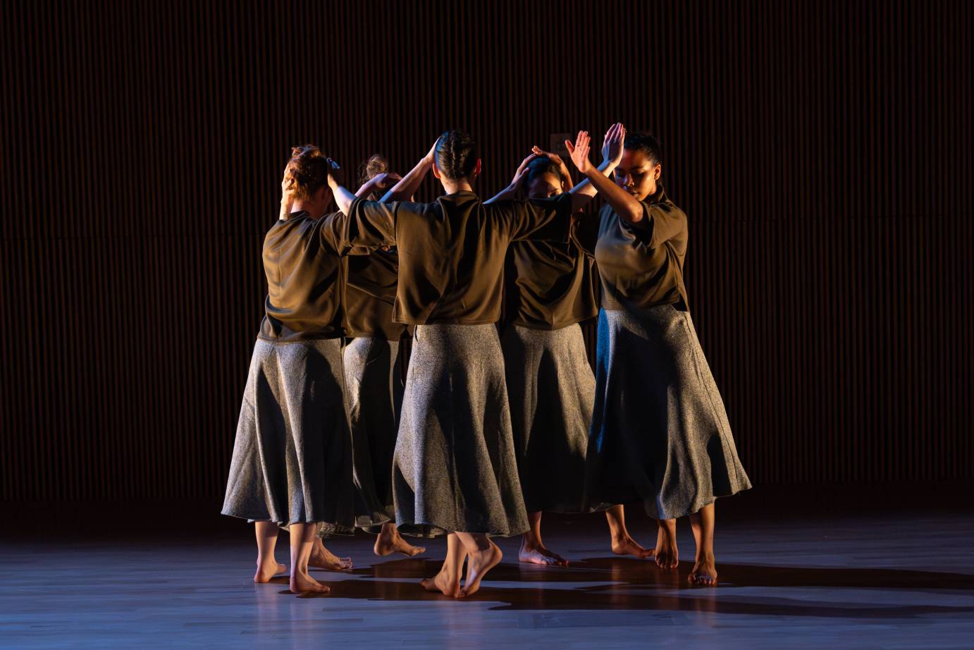 Standing women dancers in a circle face one another dressed in brown tops and silvery calf-length skirts. Their wrists cross the dancer on either side of them, face height.