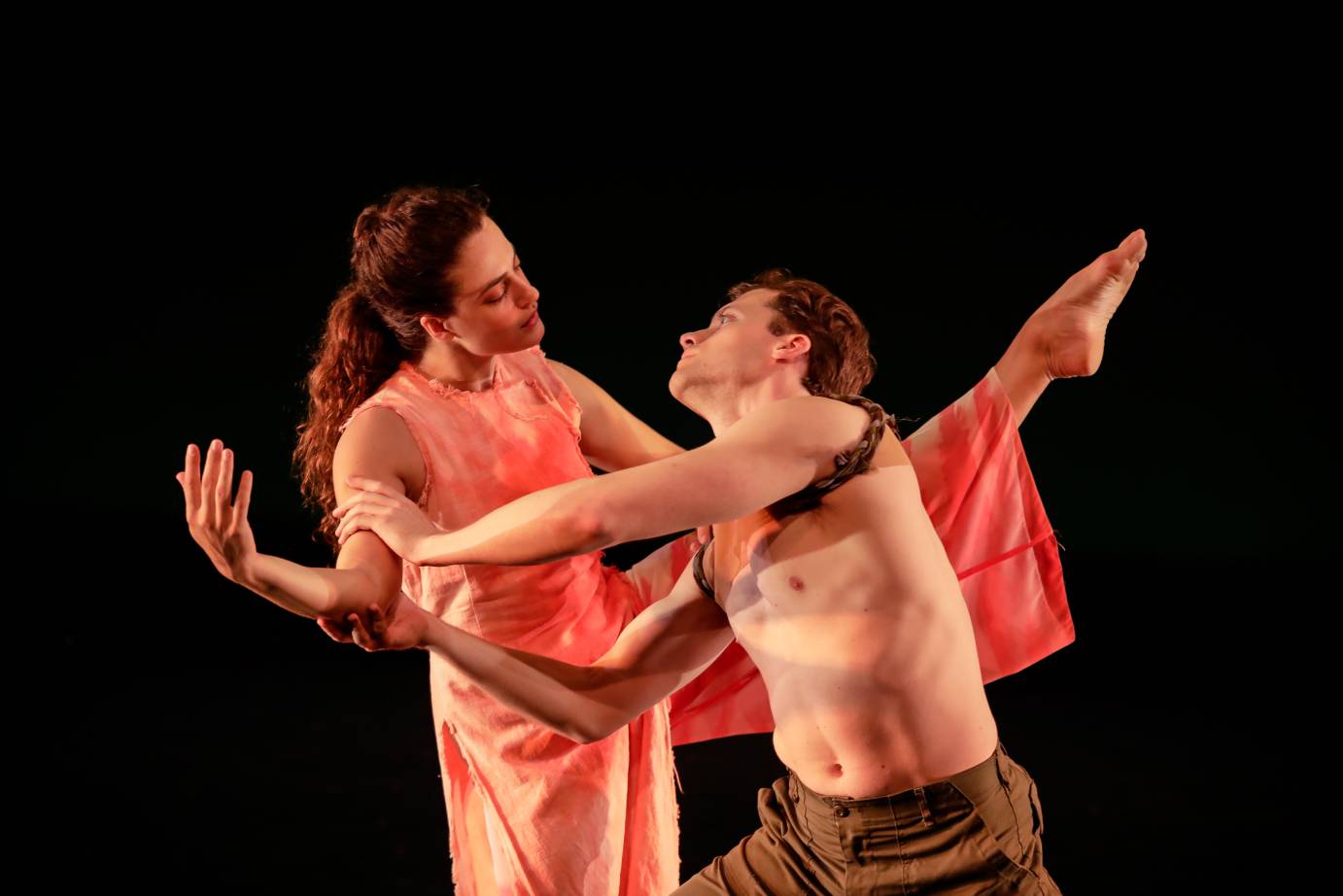 A woman and a man, she in an salmon-colored costume and he with a fabric ringing his shoulders, face one another tenderly as they slightly lean away from one another. He clasps her upper right arm and she lifts her leg.