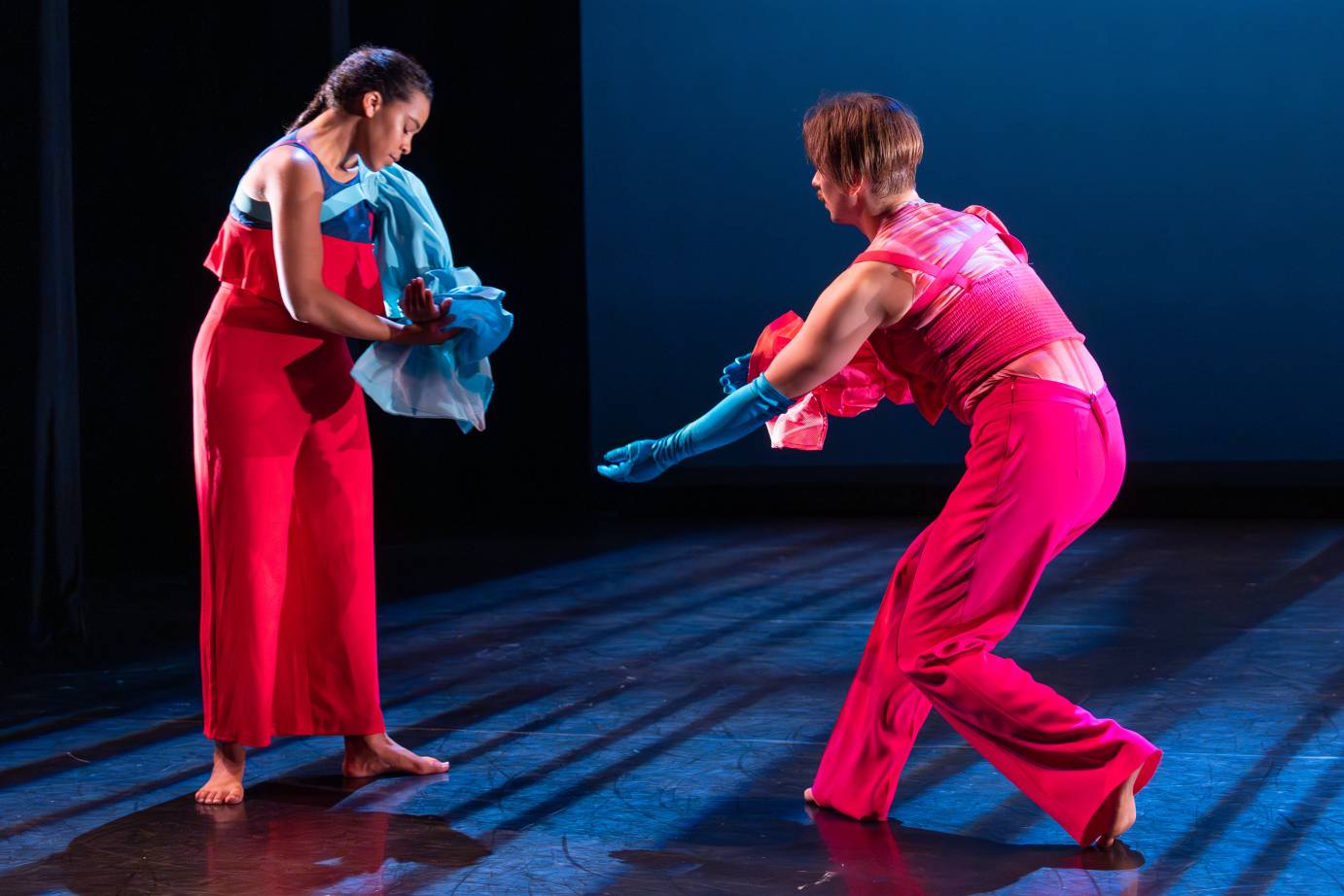 A woman and man face one another dressed in red and blue. The woman looks diagonally down while the man reaches with a blue gloved hand.