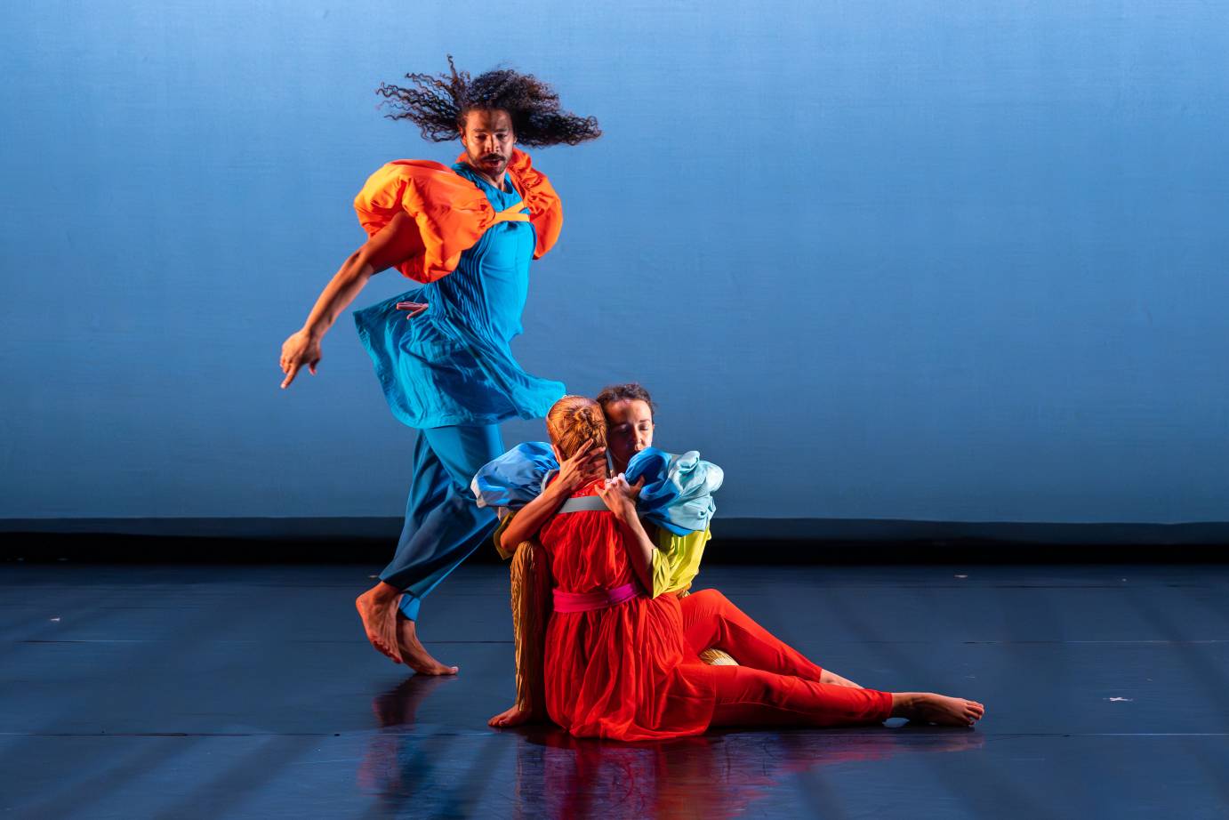 A woman costumed in a red dress, seated with her back to the audience, embraces a man in blue and yellow. A skipping man, in a neck to ankle costume adorned with orange puffs around the upper arm, looks astonished as he watches the couple.