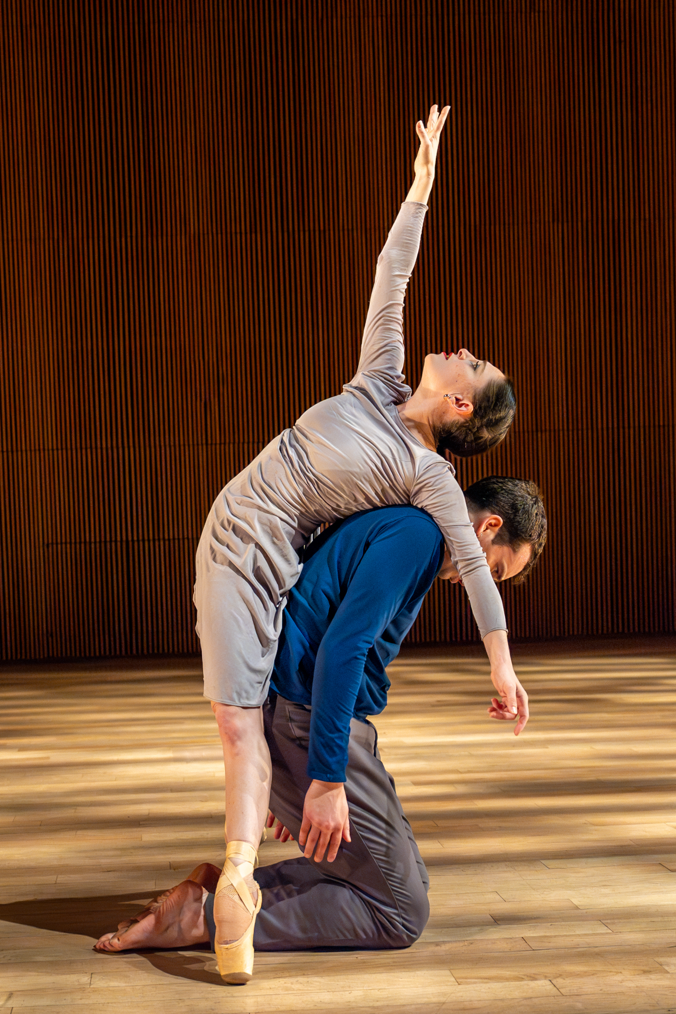 A woman in a form-fitting gray dress leans back over a crouched man, her arm and gaze upward.