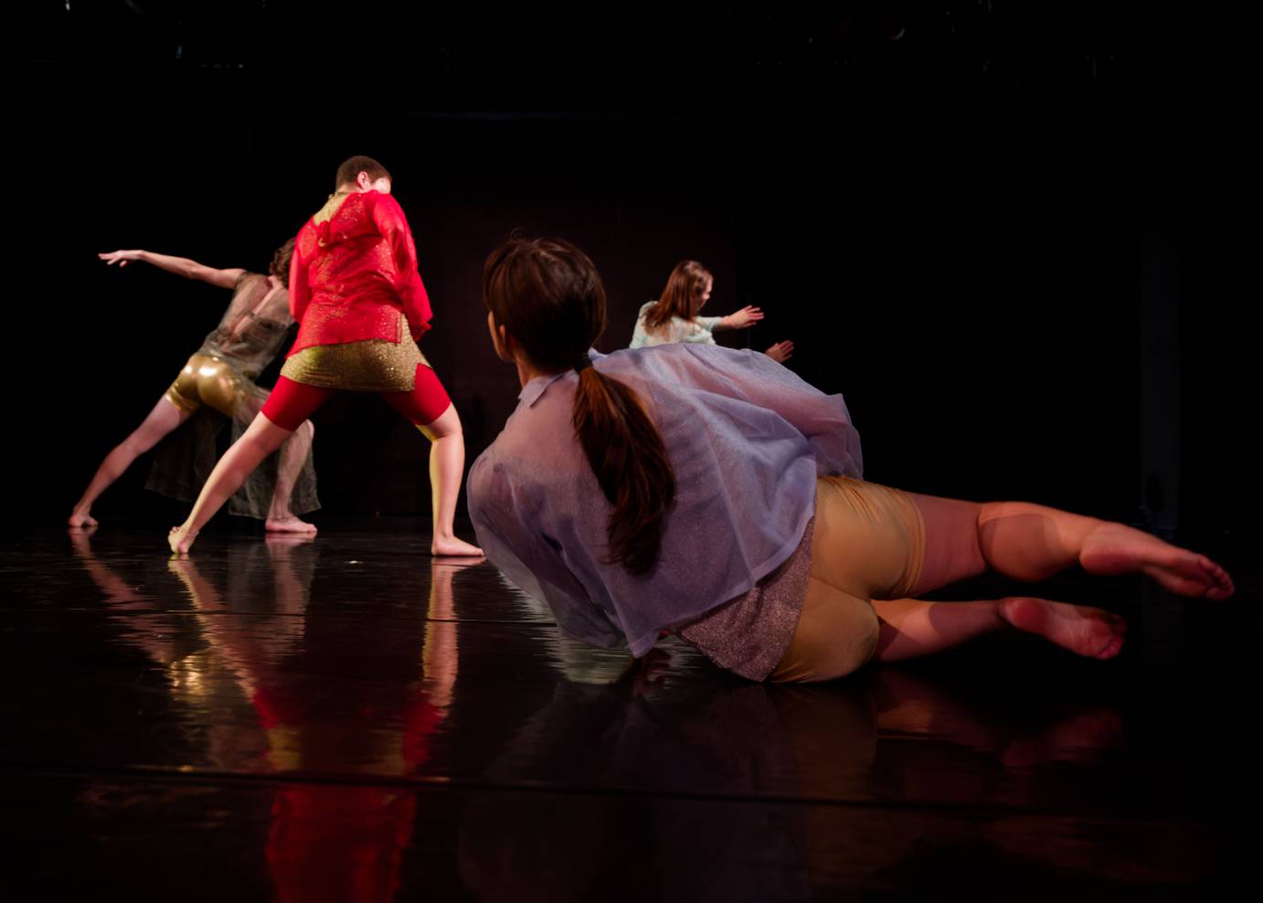Four dancers - the long, dark pony-tailed woman on her side on the floor in lavendar top with her back to the audience - contrasts with the three others who standing dressed in gold, red, and seafoam.