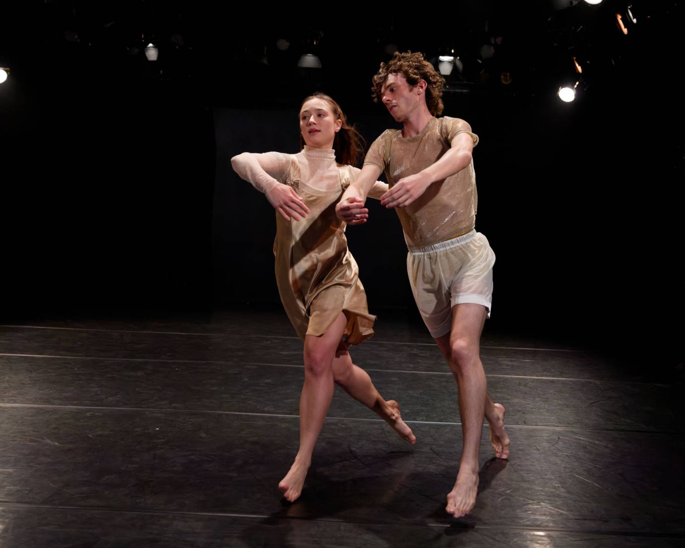 A man and woman running with arms circled in front of their bodies, tilt toward their right. They are dressed in gold flecked tops. He in white shorts.