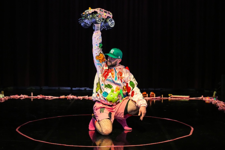 a bearded figure encircled by toys wearing a bright green baseball cap and a colorfully designed wind breaker raises holds a pink big wheeled toy truck above their head.
