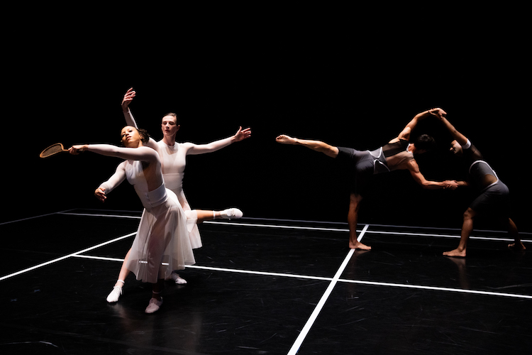 dancers dressed in gauzy modern versions of tennis outfits some in white some in black cavort on a stage decorated by neat strips of white tape.