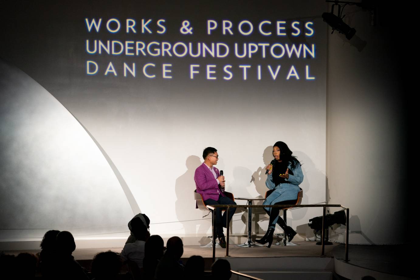 A moderator dressed in an aubergine colored jacket holds a mic while speaking with the choreographer dressed in blue with long, black hair.