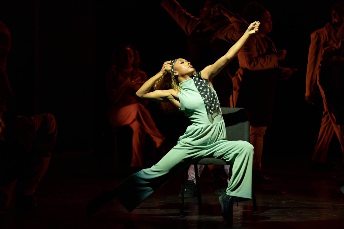 A woman seated in a chair wearing a bluegreen jumpsuit, lifts their face, chest and arm upward while their right leg reaches back. The background is dark.