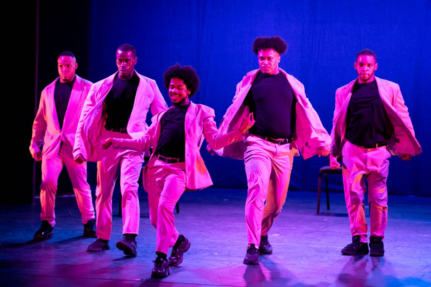 Five male performers bathed in purplish light, face the audience wearing suit jackets, pants, and belts. The performer in the foreground smiles as their arms gesture at their sides. The performers behind express serious faces.  