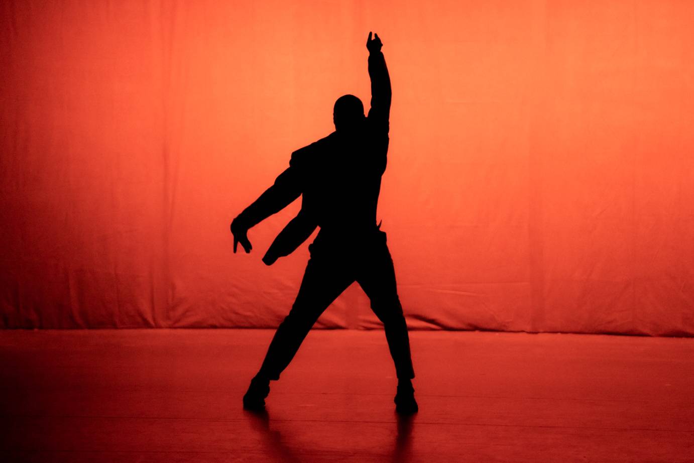 A lone performer against an orange background in silhouette lifts one arm upward. 