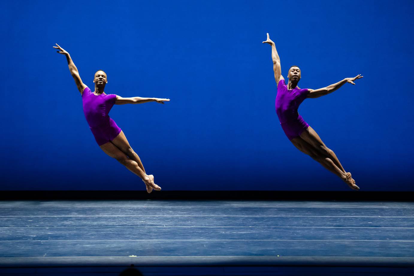 Two men in purple leotards and tights jumping in arabesque against a black screen