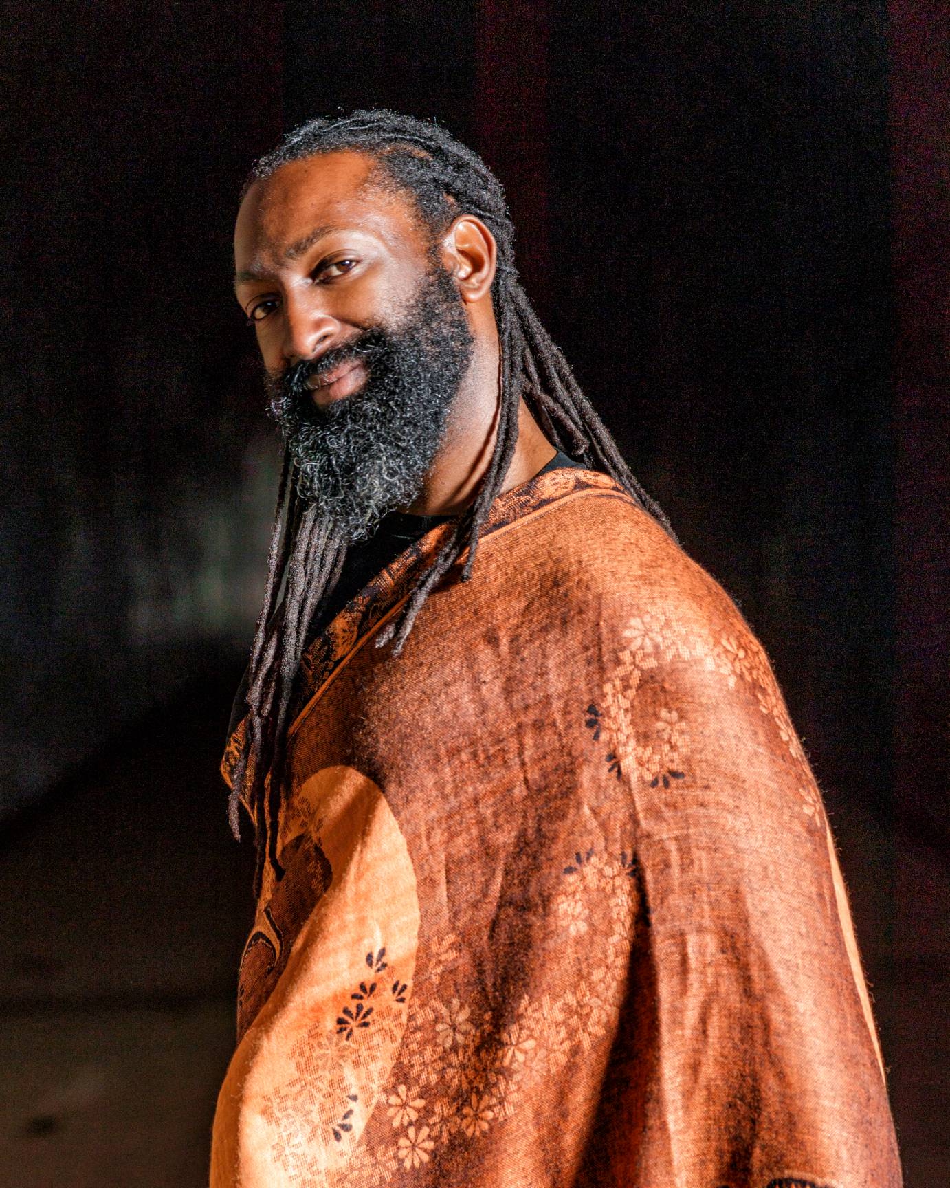 A bearded man with long braids looks to the camera with head tilted, wearing a leather-like poncho.