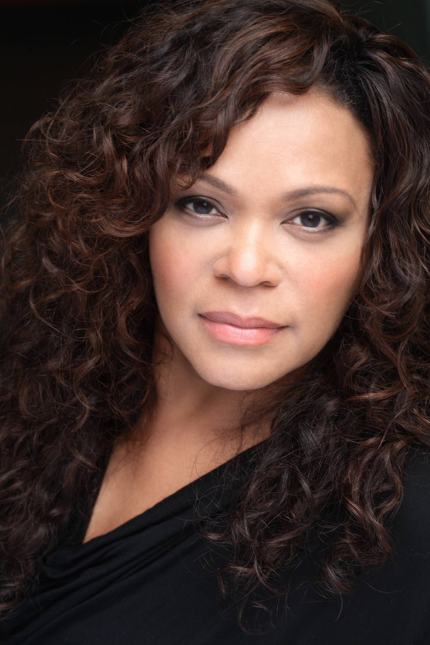 A dark-eyed woman, with peach-colored lips, and long wavy dark hair looks to the camera with an intense expression.