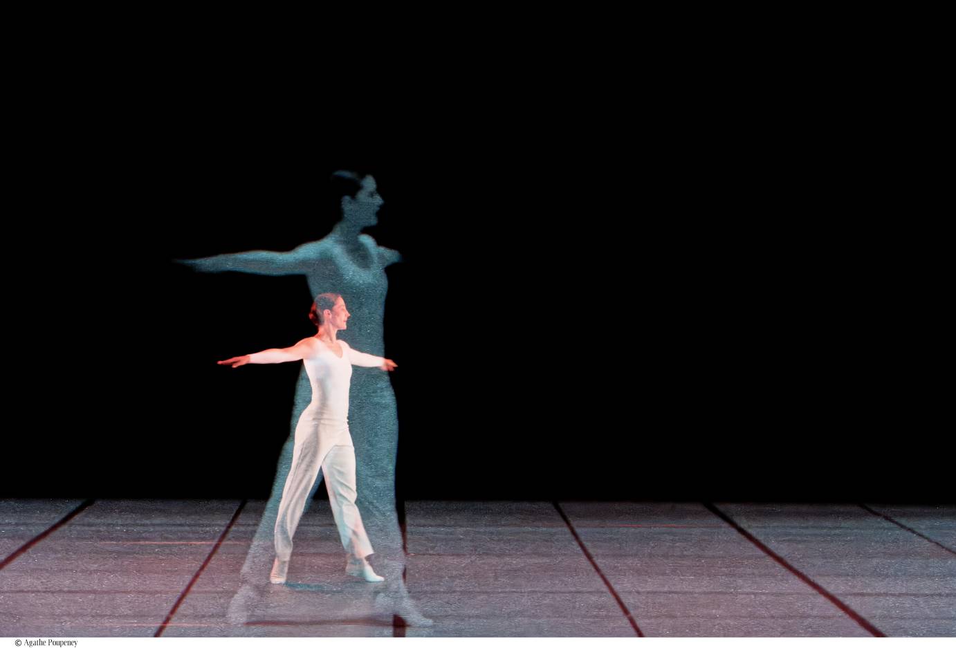 Solo woman facing stageleft, dressed in a long-sleeved white unitard, with a small bun, stands on releve, arms extended horizontally, her filmed image directly behind