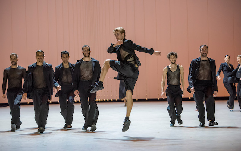 A dancer leaps into the air while a line of dancers walk behind him.