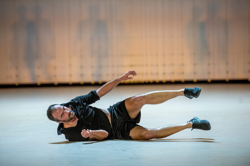 A man lays on his side in midst of rolling on the ground. He wears a button up shirt, shorts and sneakers.