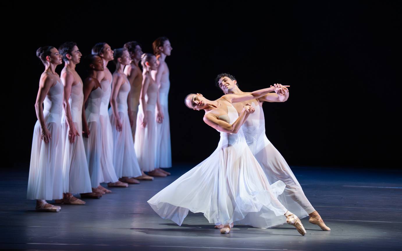 A luxurious woman and man duet bends and seemingly swings forward against a backdrop of several upright rigidly positioned ballerinas in a line.