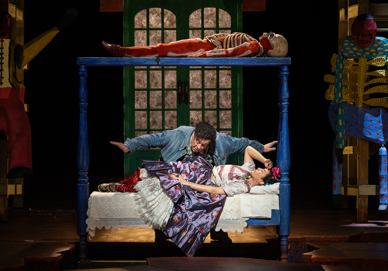 Frida lies on her little blue single bed with Diego hovering over her. A skeleton lies on top of the canopy.