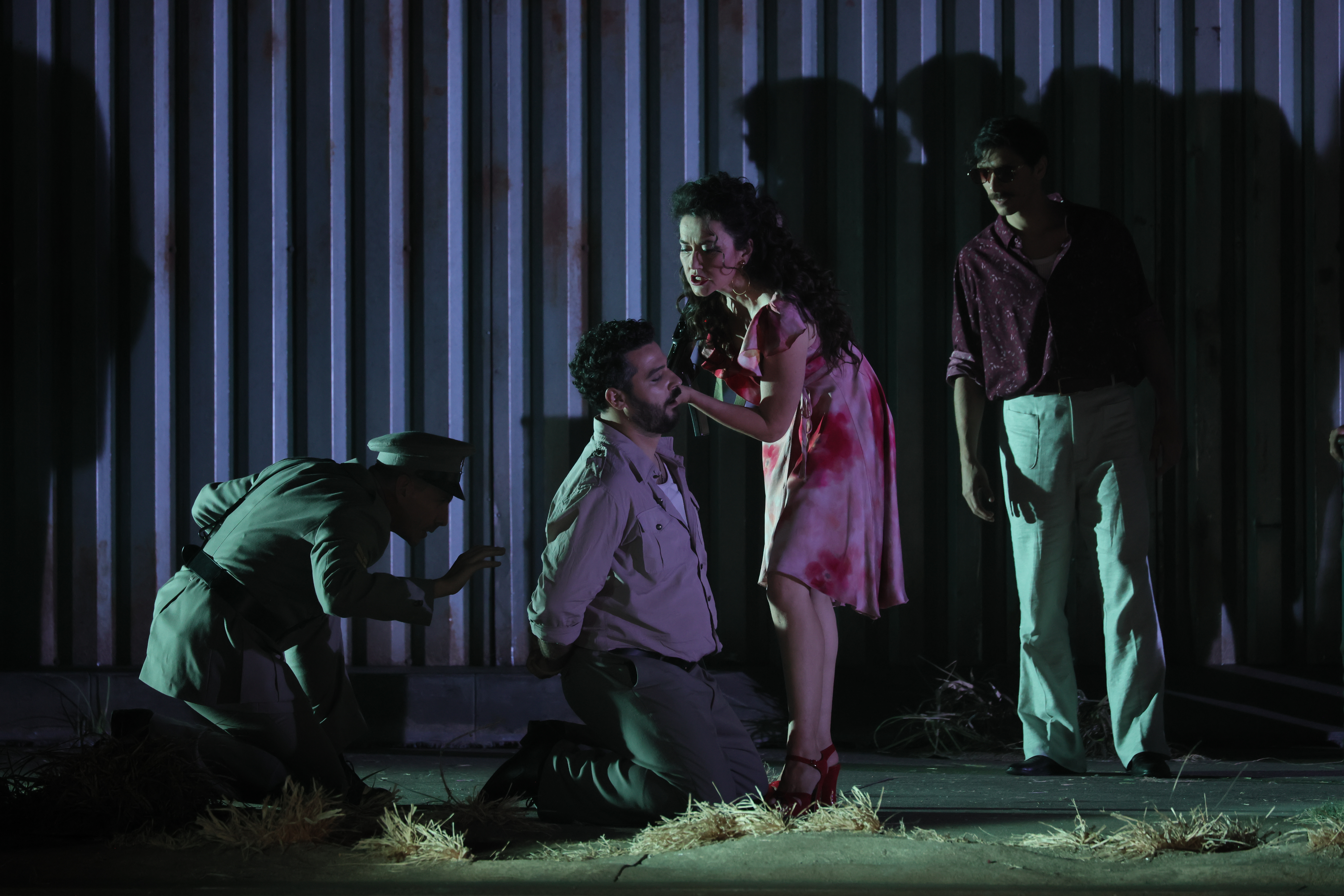Carmen, in pink flowered dress, leans close to a handcuffed man on his knees, who stares unemotionally.