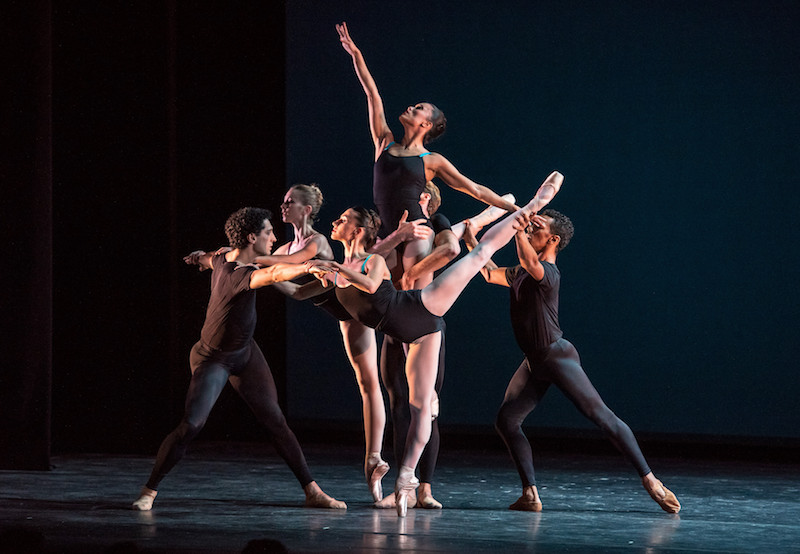 Two ballet dancers strike an arabesque pose. One is on en pointe and the other is lifted in the air. Other dancer encircle them and some lift the raised dancer above them.
