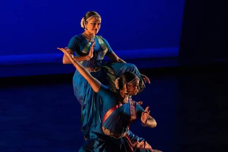 a female duet dressed in deep teal and gold gowns with jeweled hair ornamentation, strike a pose. One stands, peering at the audience with a piquant expression  and the other sits with her face in profile, more serious in disposition. 