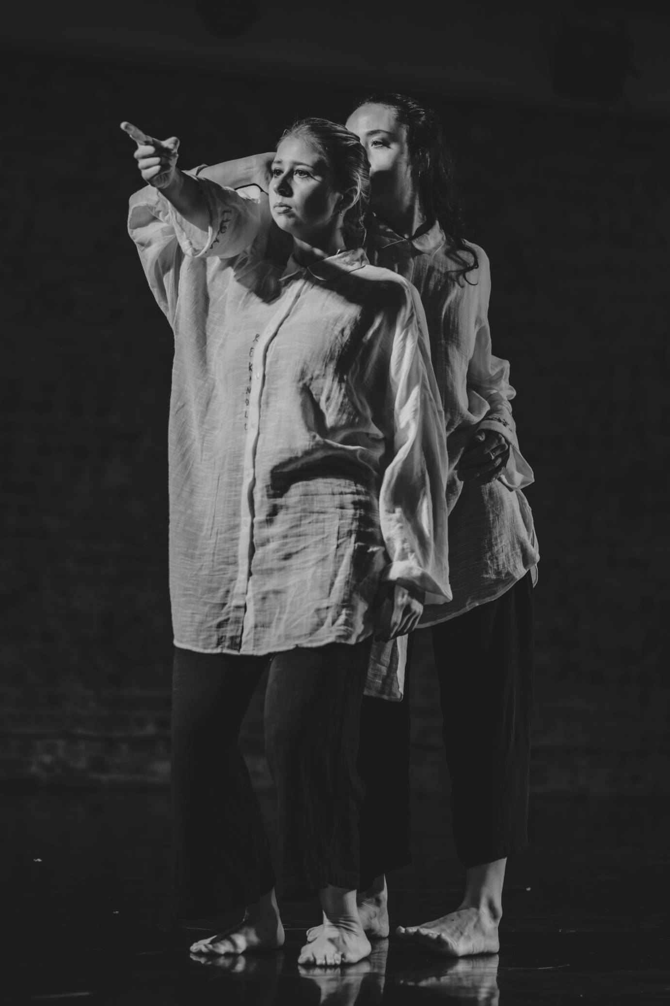 A black and white photo featuring two women one behind the other. The dancer in back points through the bended arm of the dancer in front. Both look toward the diagonal.