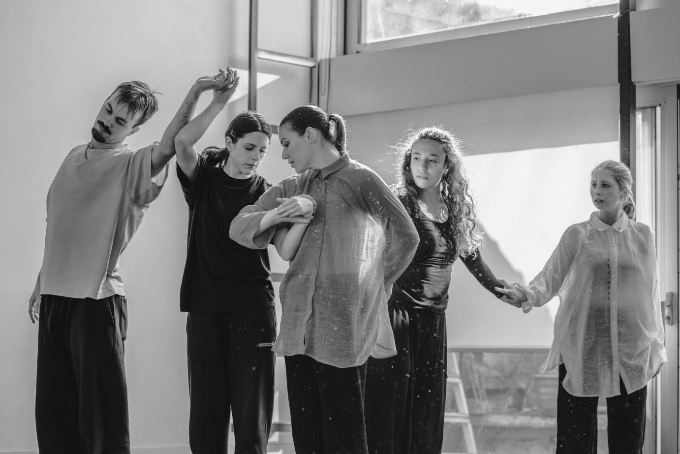 A black and white photo featuring one man and four women who hold hands above their heads or at their waists. There is an easeful and elegant quality to their relationship.